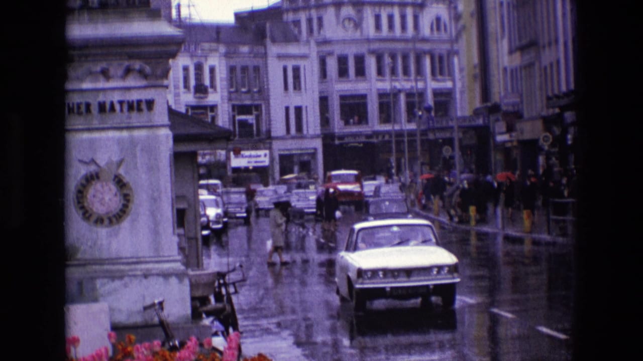 1969 Ireland street view with wet pavement, classic cars, pedestrians under umbrellas, and shopfronts in the background.
