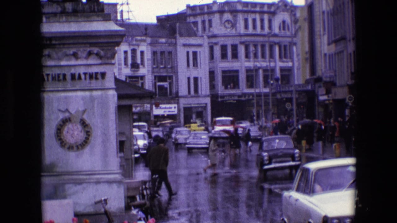 Rainy 1969 Ireland street scene showing pedestrians, umbrellas, vintage cars, and commercial buildings behind a stone column.