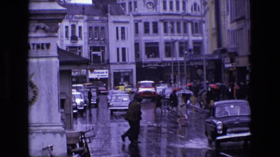 1969 Ireland street view with a pedestrian crossing in rain, vintage cars, umbrellas, storefront signs, and reflective pavement.