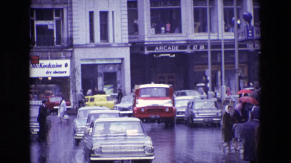 1969 Ireland commercial street with rainy traffic, a red van, classic cars, pedestrians with umbrellas, and an Arcade sign.