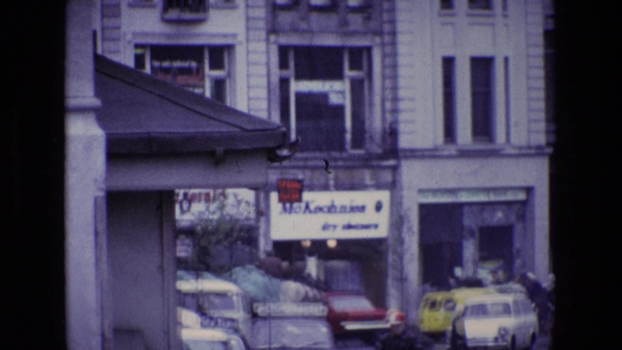 Rainy town street with shopfronts, parked cars, pedestrians and a dry cleaners sign.