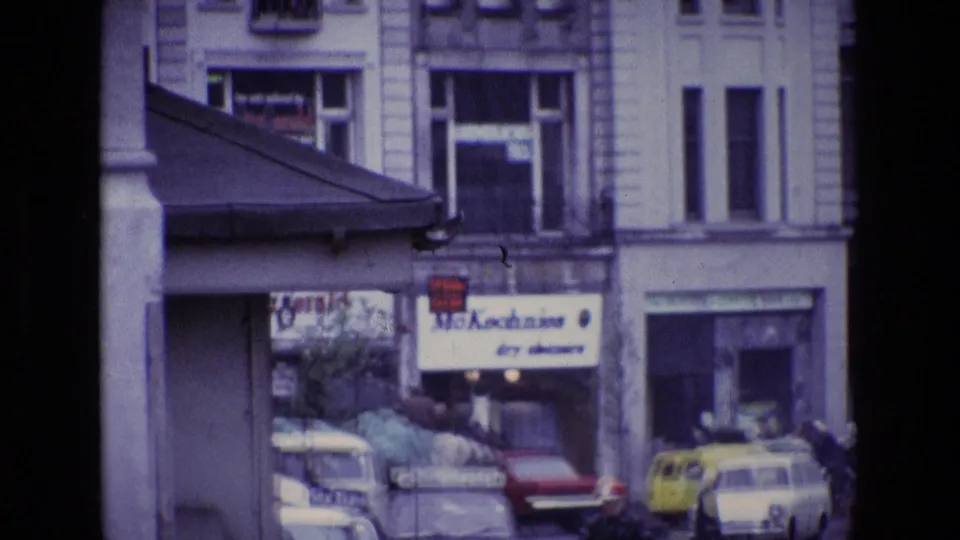 1969 Ireland street view showing wet pavement, commercial buildings, classic cars and pedestrians near a dry cleaners sign.