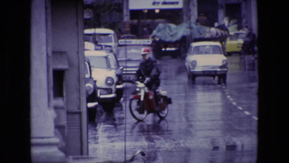 1969 Ireland rainy street view with a motorcyclist in the foreground, vintage cars, pedestrians and shop signage.