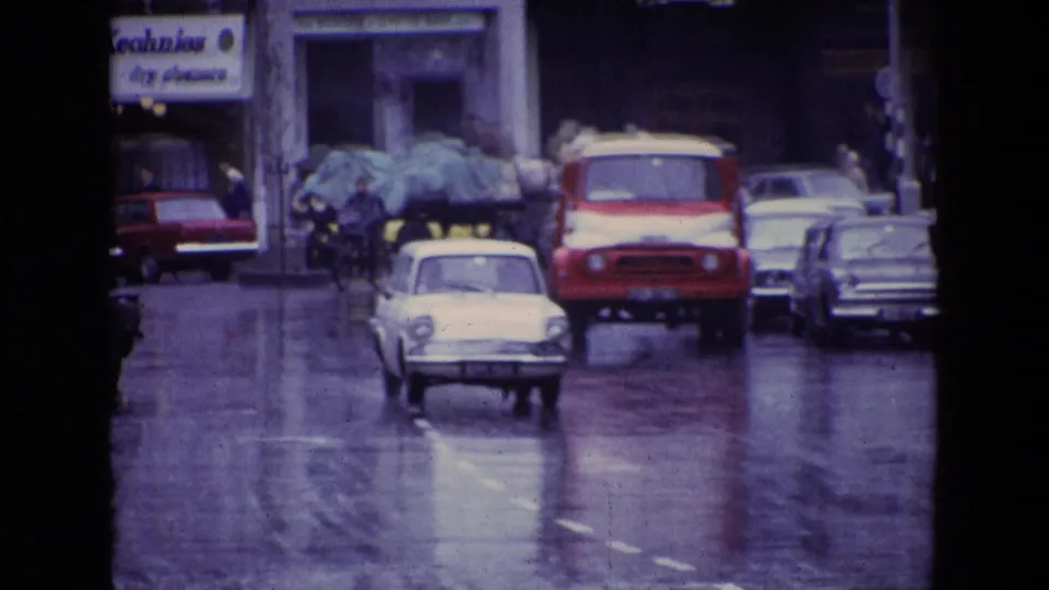 1969 Ireland town traffic scene showing wet pavement, compact cars, a red-and-white truck, pedestrians and commercial signage.
