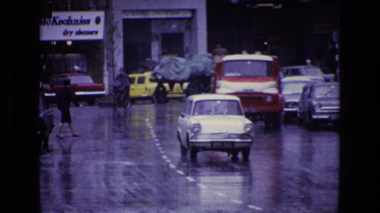 Rainy city street with pedestrians, 1960s cars, and a red-and-white van on wet pavement.