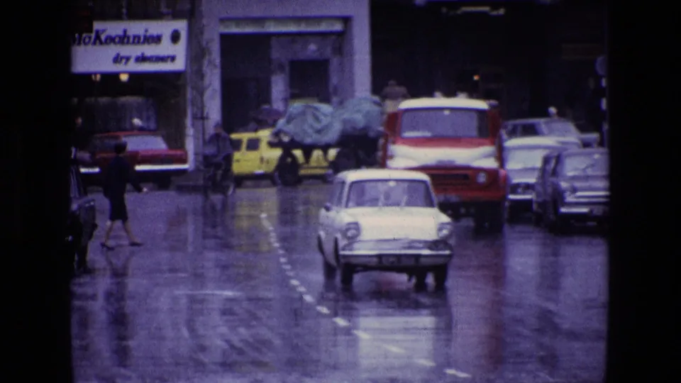 1969 Ireland street scene with wet roads, pedestrians, compact cars, and a red-and-white van moving through rain.