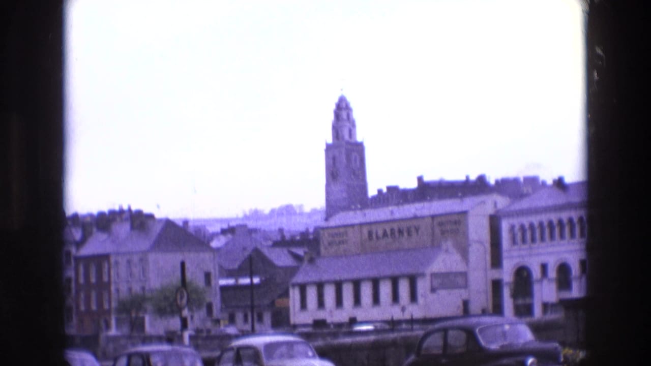 Clock tower above rooftops with parked cars and a large BLARNEY sign on a building.
