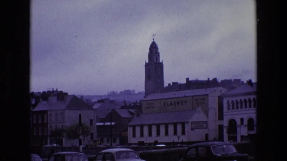 1969 Ireland town view with clock tower rising behind low buildings, parked cars, and the BLARNEY sign in rainy light.
