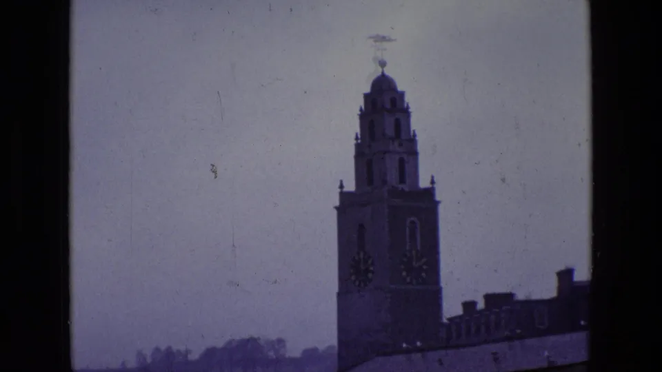 1969 Ireland 8mm frame showing a distant clock tower rising above a roofline under a muted overcast sky.