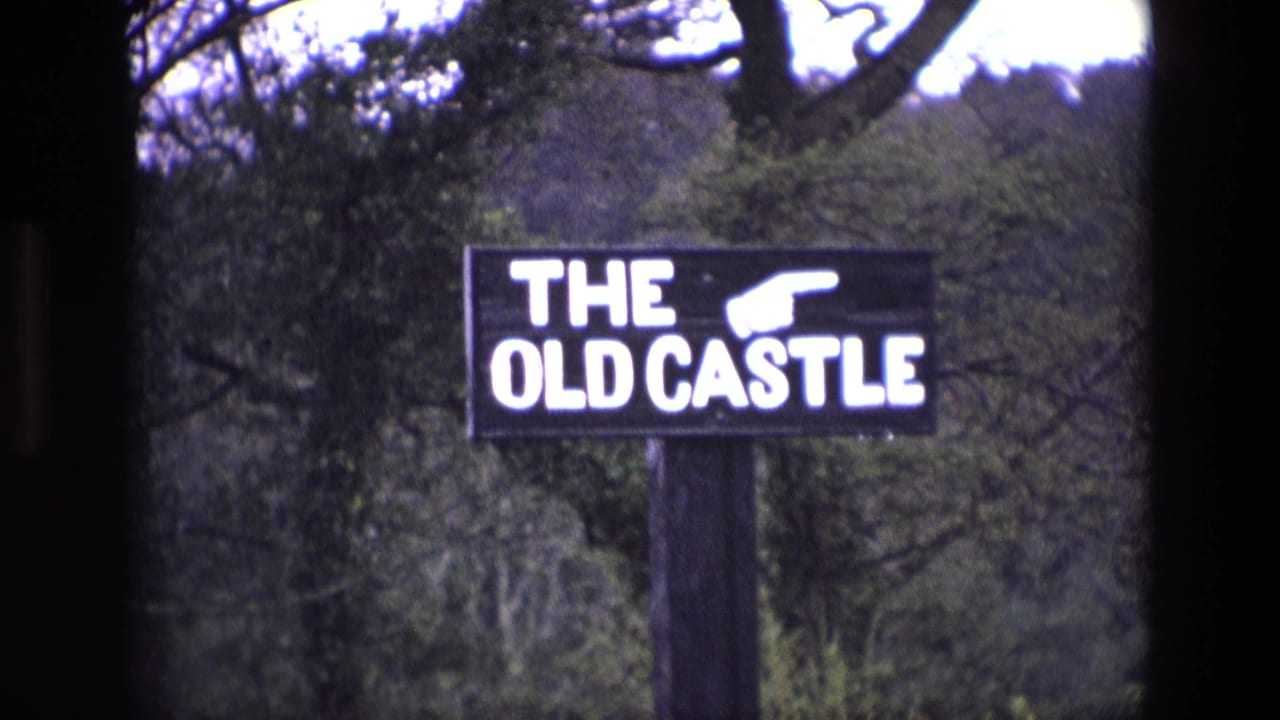 1969 Ireland 8mm frame of a black roadside sign reading THE OLD CASTLE with a white pointing hand, set before trees.