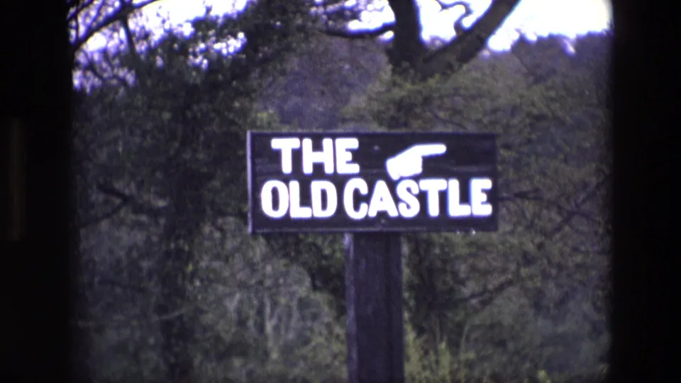 1969 Ireland 8mm frame of a black roadside sign reading THE OLD CASTLE with a white pointing hand, set before trees.