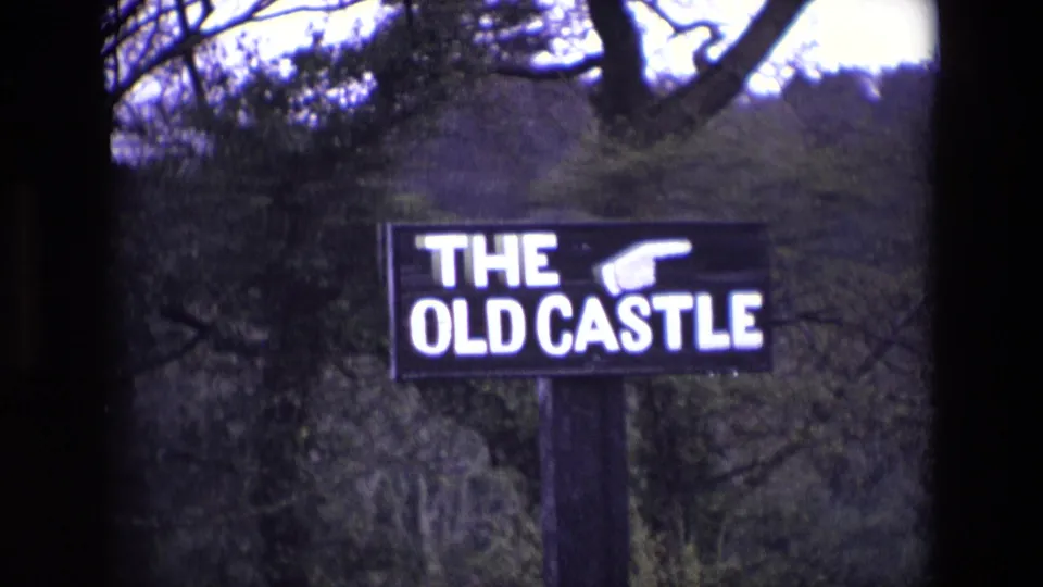 1969 Ireland 8mm frame showing a closer view of the THE OLD CASTLE sign, its white letters and pointing hand visible against woodland.