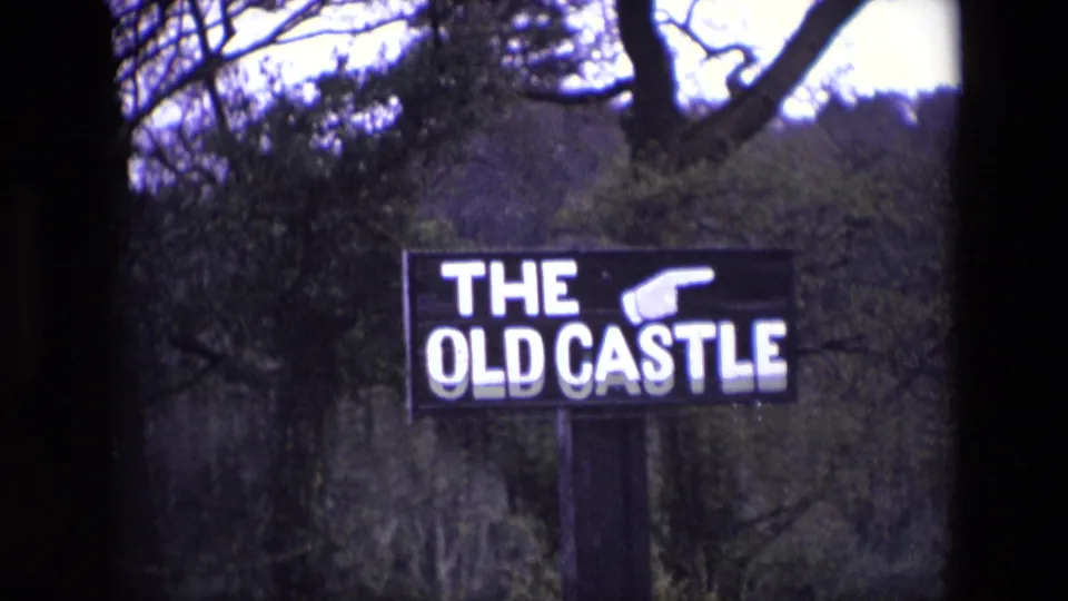1969 Ireland 8mm frame with the THE OLD CASTLE sign on a post, white pointing hand and lettering visible among trees.