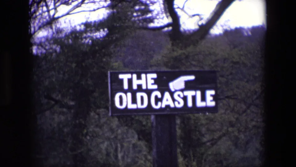 1969 Ireland 8mm frame showing a black THE OLD CASTLE sign with a pointing hand in wooded grounds.