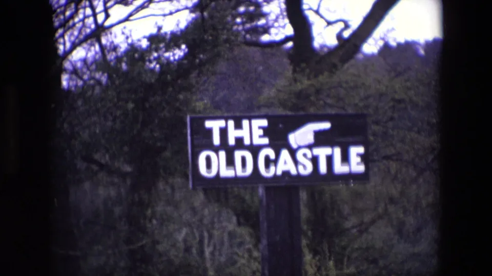 1969 Ireland home-movie frame of the THE OLD CASTLE sign, partly framed by dark film edges and trees.
