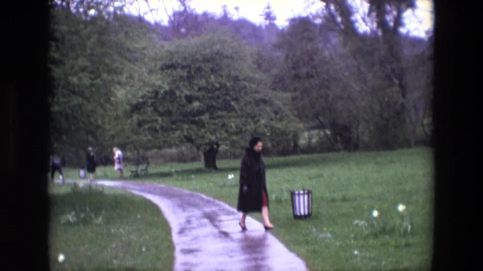 1969 Ireland 8mm frame with a woman walking near a wet path, lawn, trees, and distant visitors in a park-like setting.
