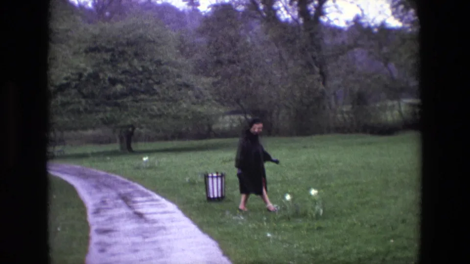 1969 Ireland home-movie frame showing a woman in a dark coat crossing grass beside a wet path in wooded grounds.