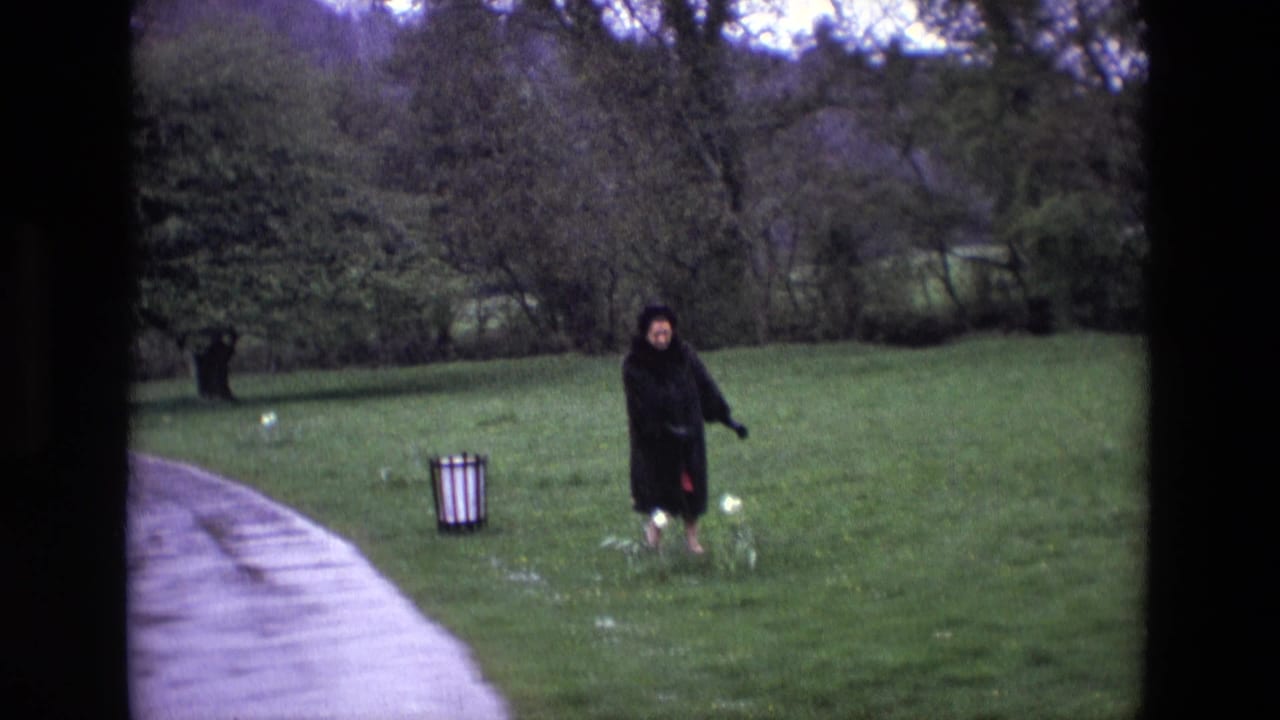 Woman in a dark coat walks from a paved path into a grassy field near white flowers.