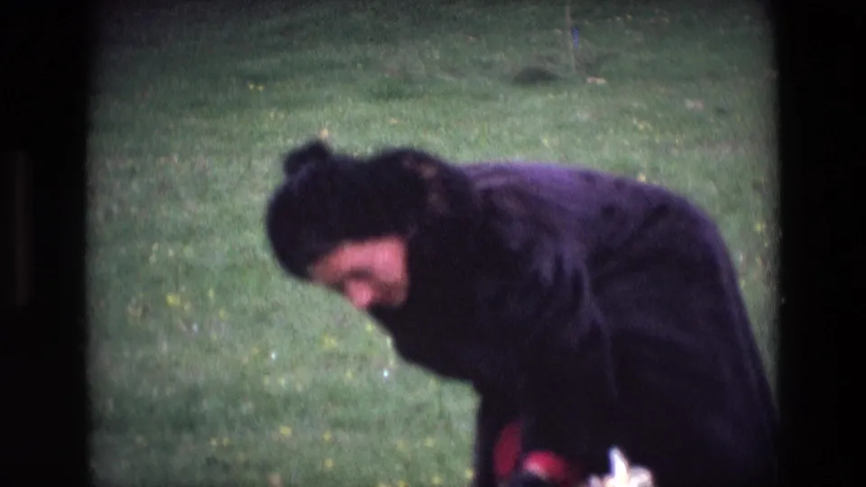 In 1969 Ireland 8mm footage, a woman in a dark coat and hat bends toward the grass in a green park.