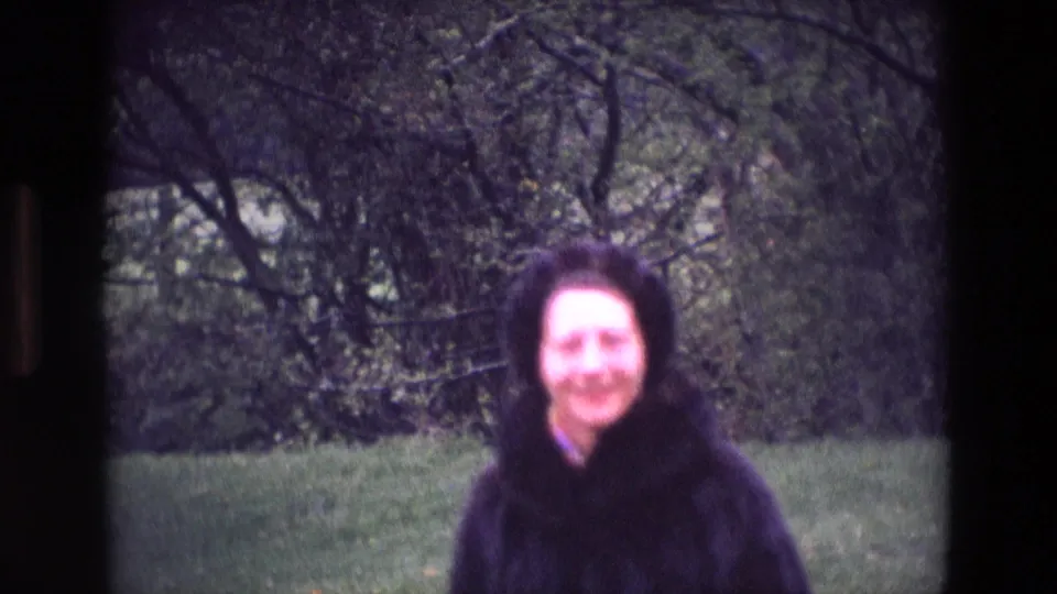 A smiling woman in a dark coat stands before trees and grass in this Ireland 8mm home-movie frame.