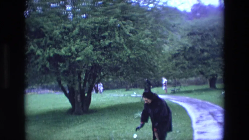 The woman bends toward the grass beside the park path, appearing to feed birds or small animals in the 1969 Irish home-movie footage.
