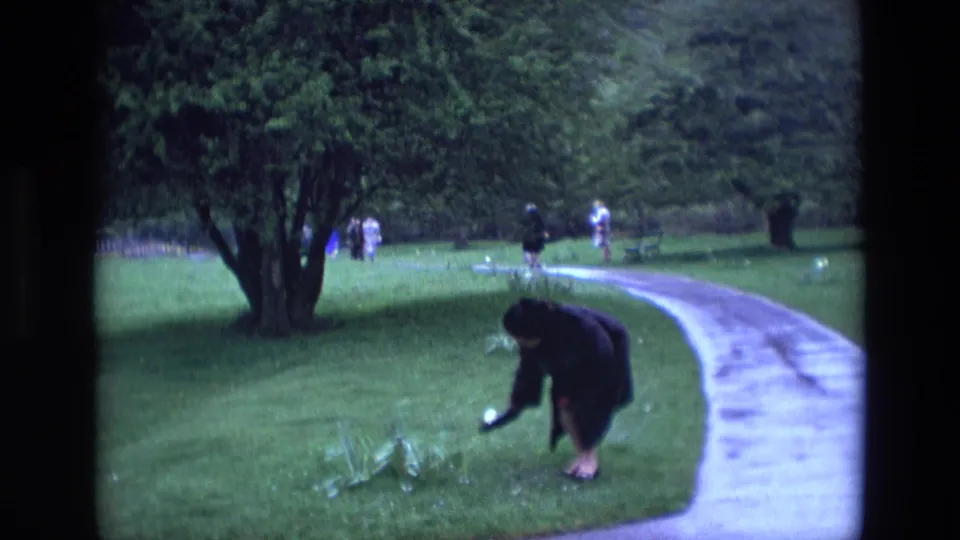 A woman in a dark coat crouches near the grass beside a curving path, with mature trees and distant park visitors behind her.