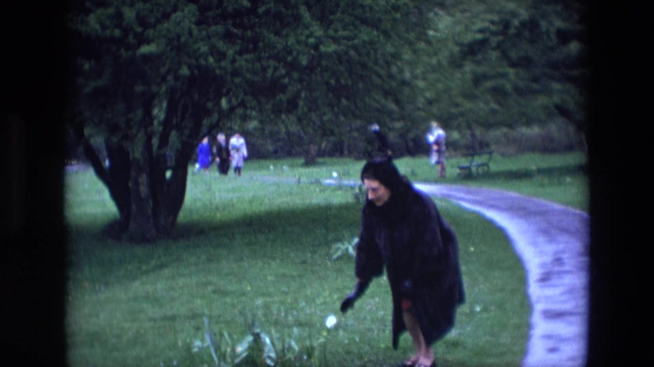 Woman in a dark fur coat bends toward flowers beside a paved park path.