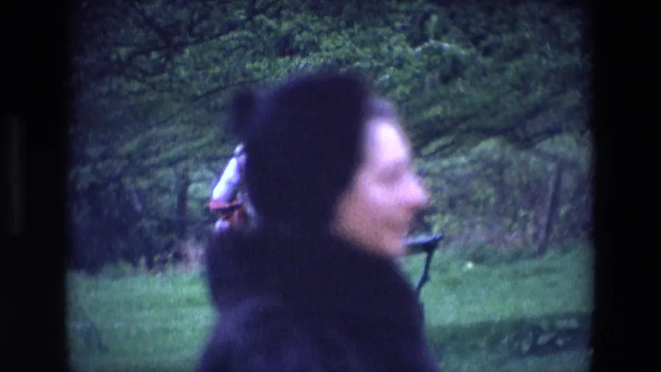 A soft, close 8mm frame shows the woman moving through the park, with trees, grass, and a bench behind her.