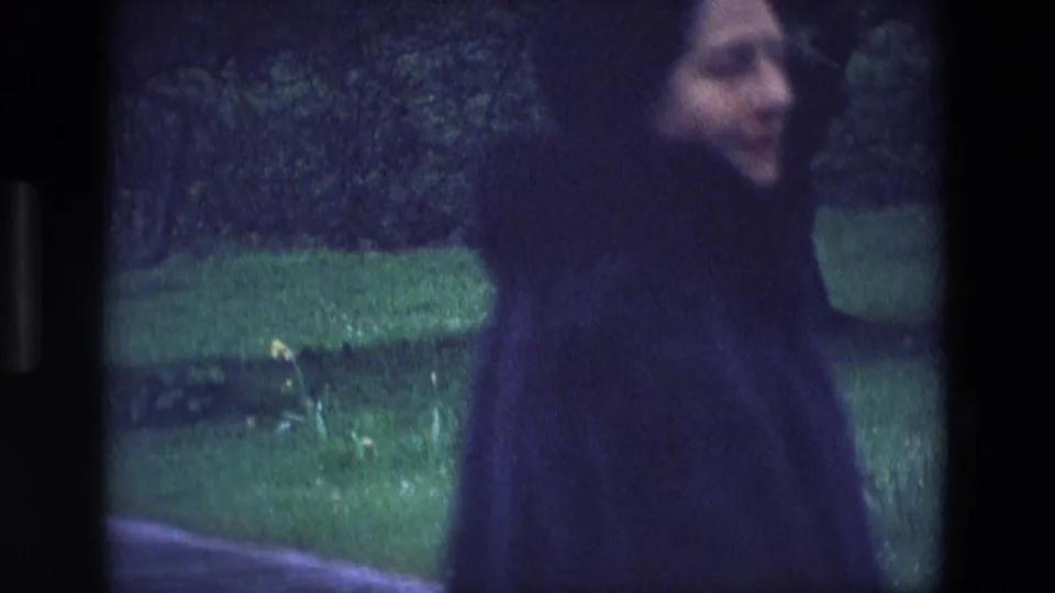The woman walks along the paved path in a green Irish park, seen in soft focus near shrubs and lawn.