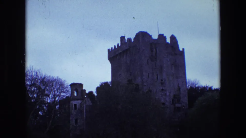 1969 Ireland home-movie frame of a shadowed stone castle and smaller ruined structure beneath a pale blue sky.