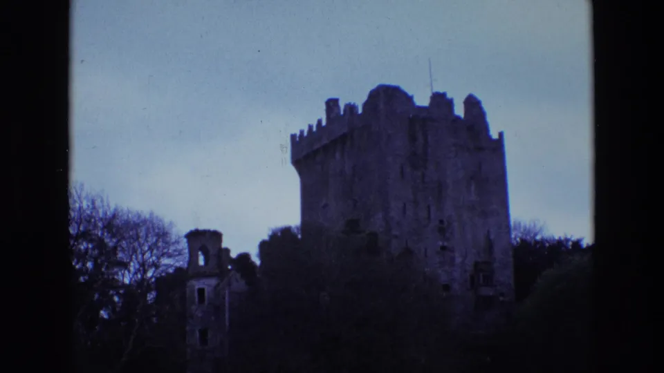 1969 Ireland 8mm frame with a dark crenellated castle tower rising behind trees, shrubs, and a smaller ruin.