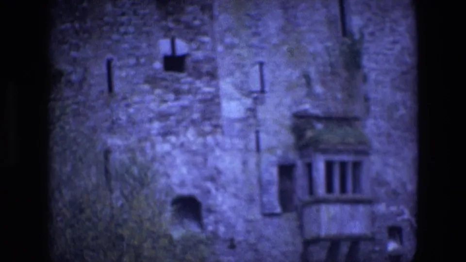 1969 Ireland 8mm frame showing a rough stone castle facade with narrow windows, a small balcony, vegetation, and dark film edges.