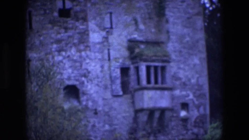 1969 Ireland 8mm frame showing a closer, grainy view of the stone castle facade, including a small balcony, dark openings, foliage, and film gate edges.