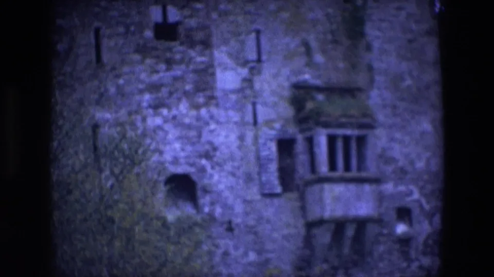 1969 Ireland 8mm frame holding on the castle facade, with narrow vertical windows, mossy stone, a small balcony, foliage, and low-light color shift.