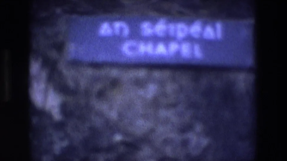 1969 Ireland home-movie frame with a blurred close-up of the blue An Séipéal Chapel sign against rough stone.