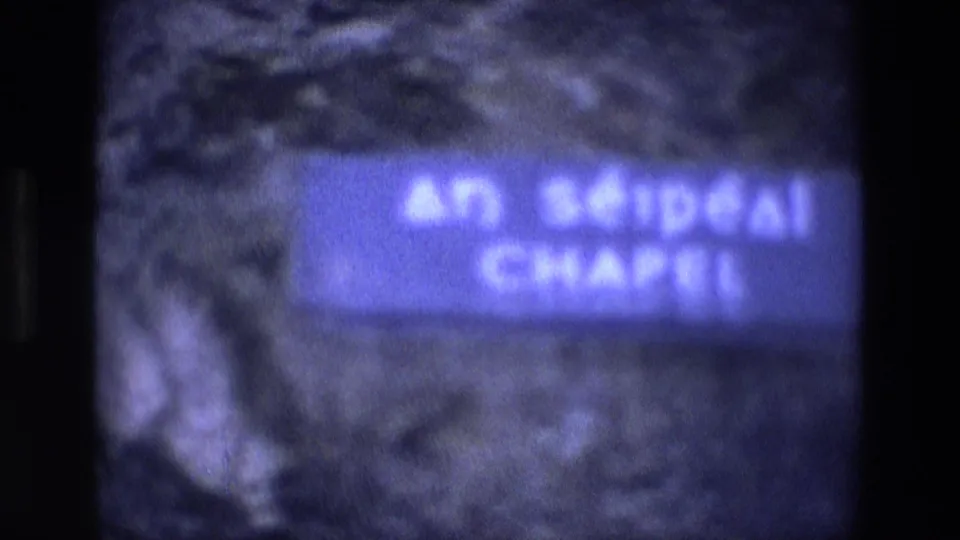 Soft-focus 8mm frame showing the blue An Séipéal Chapel sign mounted on stonework, confirming the chapel setting.