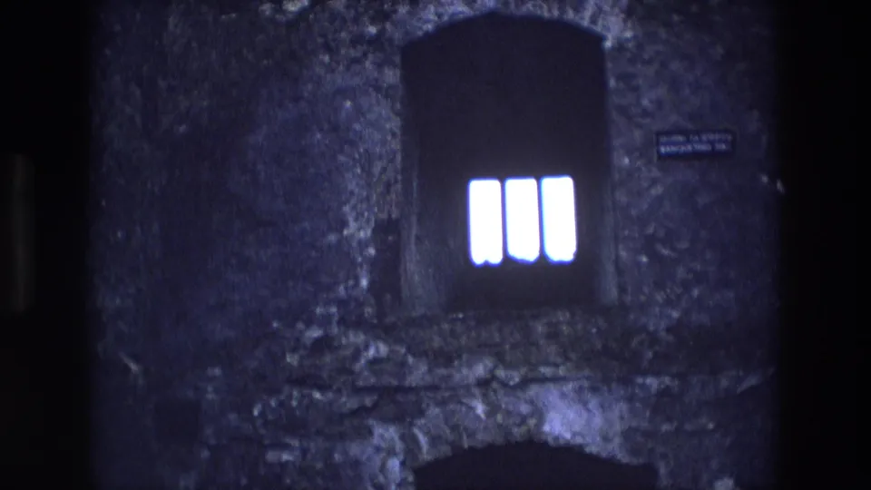 1969 Ireland 8mm frame with a dark arched window, three bright vertical panes, rough masonry, and a small wall plaque to the right.