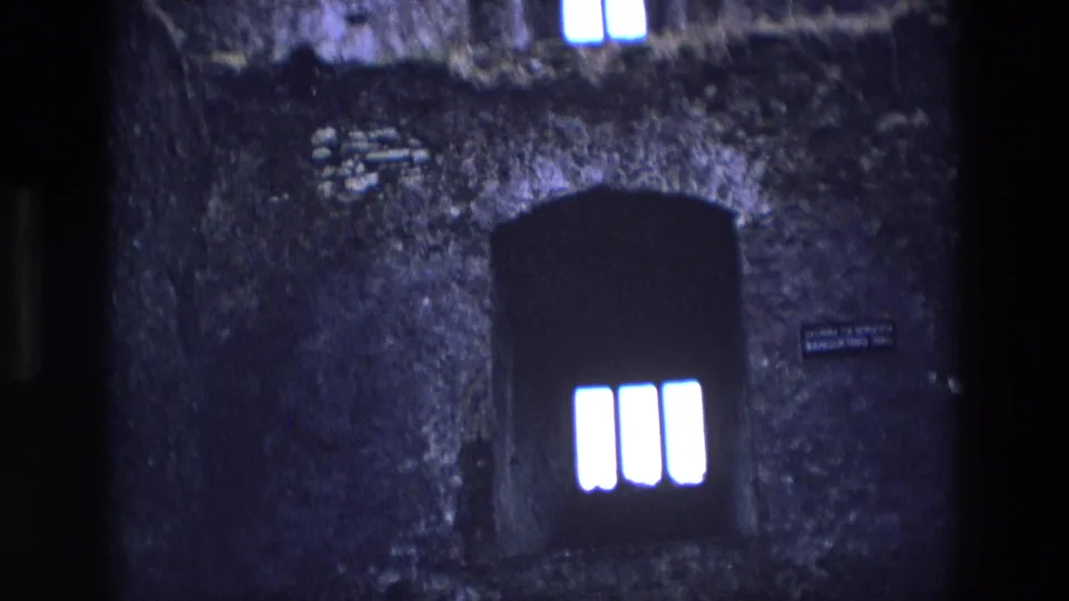 1969 Ireland 8mm frame with a dark arched window, bright barred panes, rough masonry, and small unreadable signs on the wall.