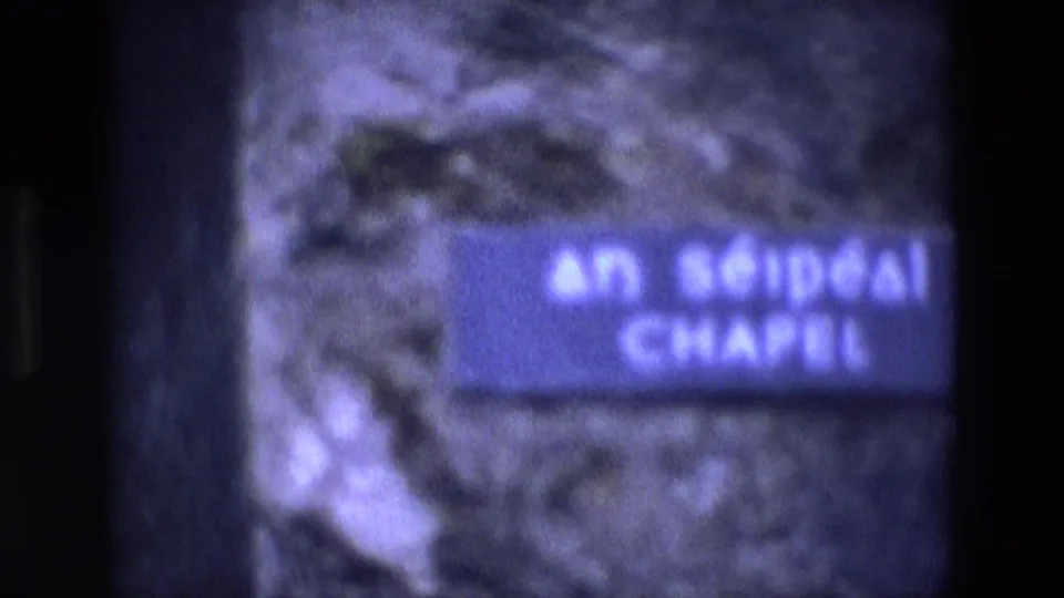 Close 1969 Ireland 8mm frame of a chapel sign reading AN SÉIPÉAL CHAPEL mounted on stone.
