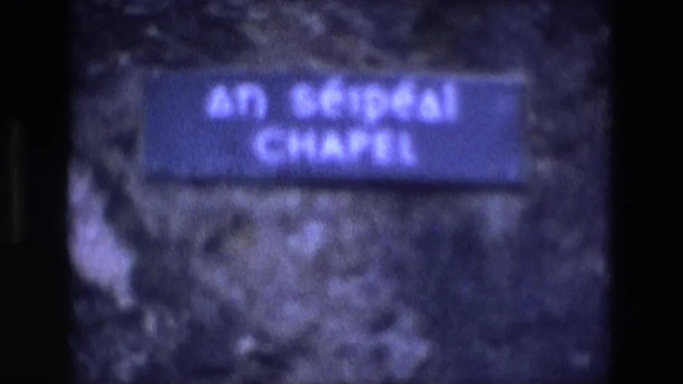 1969 Ireland 8mm frame showing a soft blue An Séipéal Chapel sign mounted near rough stone.
