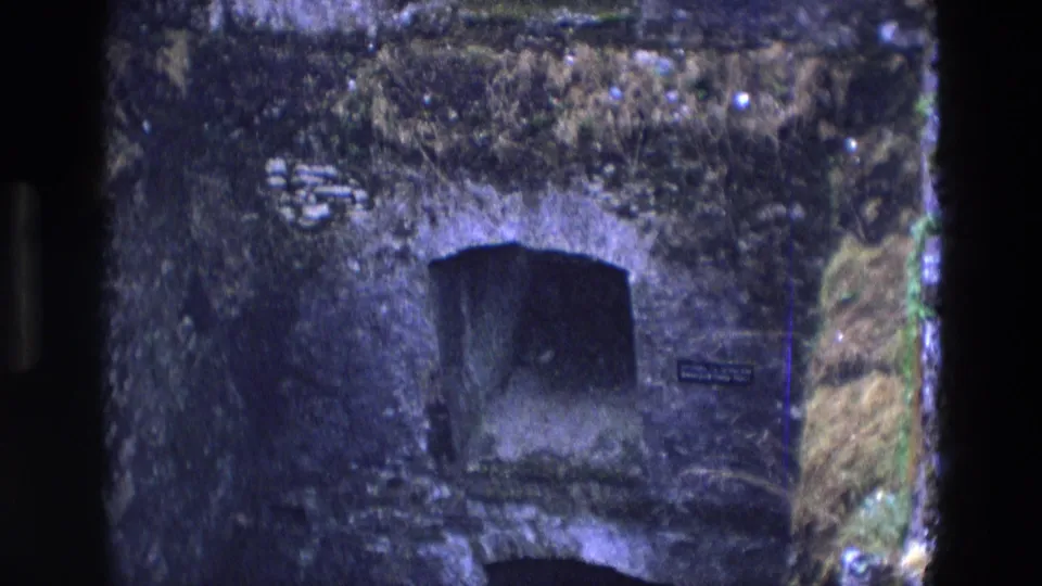 1969 Ireland 8mm frame showing a moss-covered tunnel entrance, nearby plaque, and rough vegetation around the stone face.