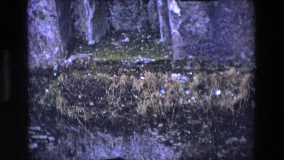 1969 Ireland 8mm frame showing the interior stone wall of an old chapel ruin with moss, rubble, and dry vegetation.