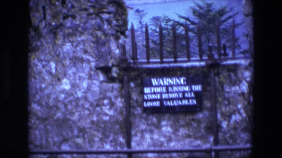 1969 Ireland frame showing an iron gate and a black warning sign about removing loose valuables before kissing the stone.