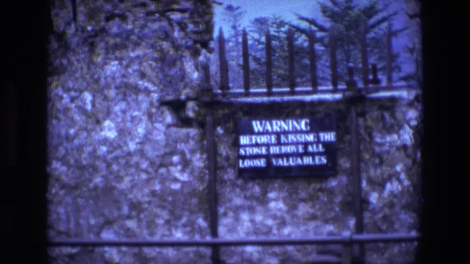 1969 Ireland 8mm frame showing a warning sign beside dark iron spikes and rough stonework at a visitor landmark.