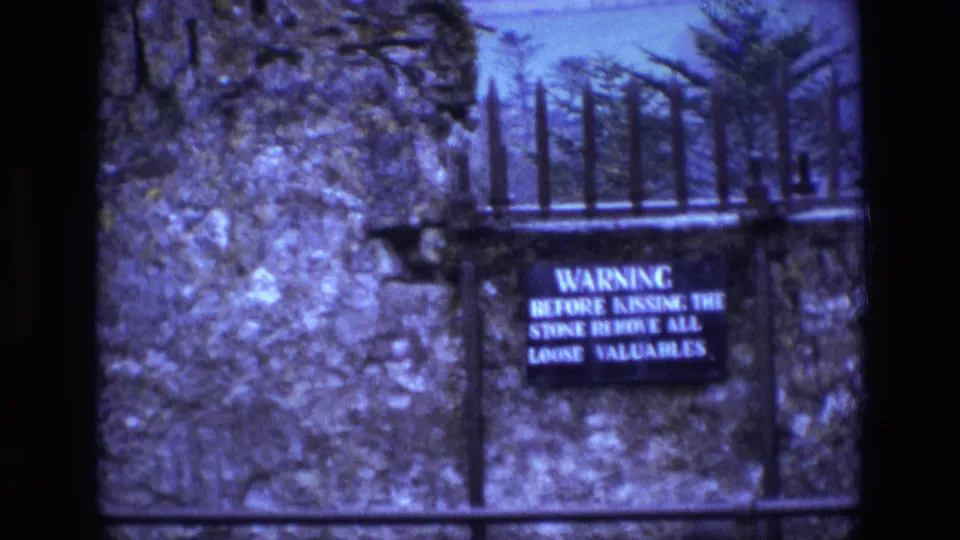 Visitor-shot 8mm view of the warning sign reading about valuables before kissing the stone, set against stone, iron spikes, and trees in Ireland.