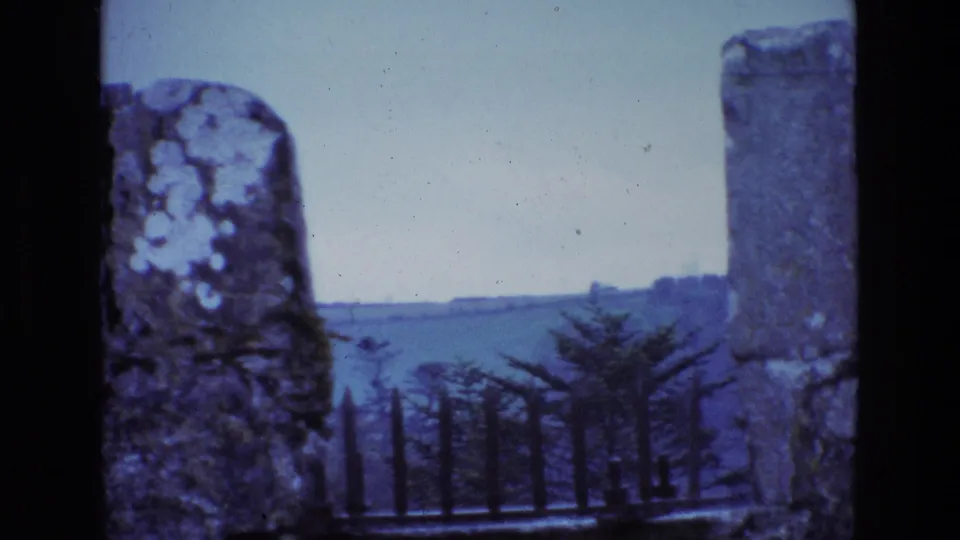 8mm Ireland landscape view framed by weathered stone posts, iron spikes, trees, rolling hills, and distant water.