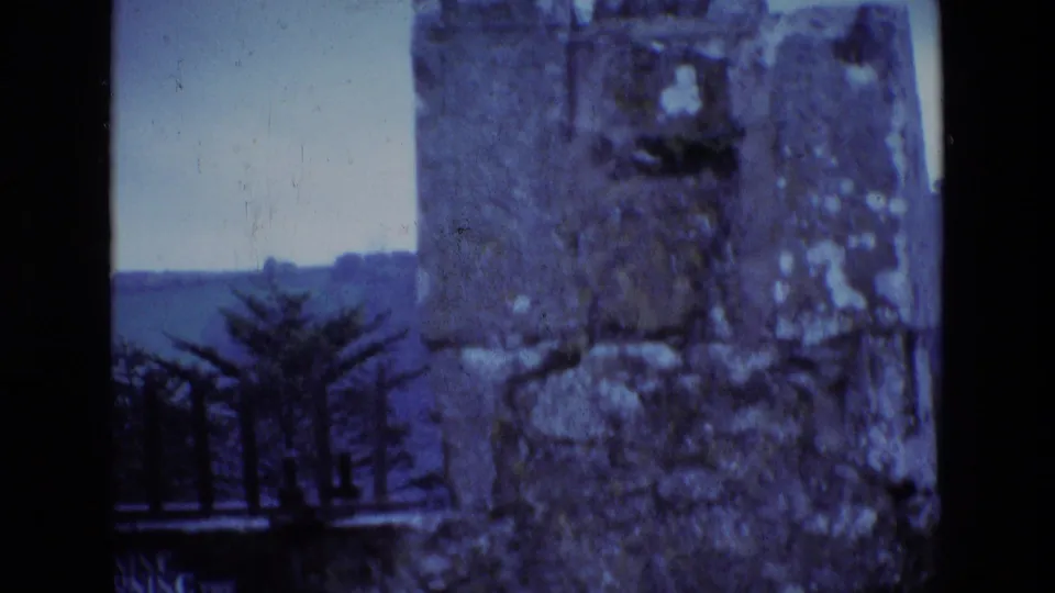 Ireland 1969 home-movie frame showing a weathered stone ruin through a dark window frame, with trees and landscape beyond.