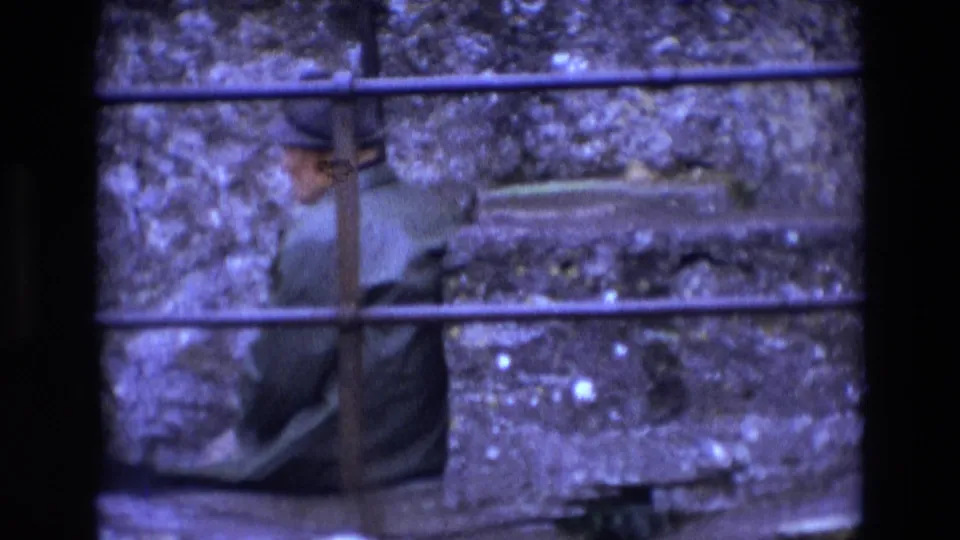 Ireland 1969 home-movie frame showing an older man in a hat and dark coat seated beside a weathered stone wall.