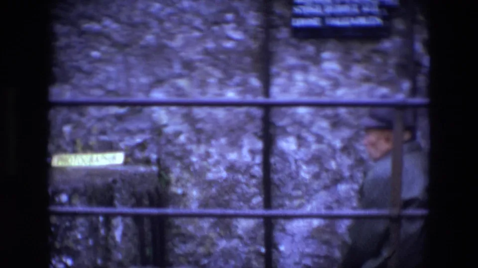 Ireland 1969 frame of an older man seated near rough stonework, seen through iron bars with a small sign in the background.