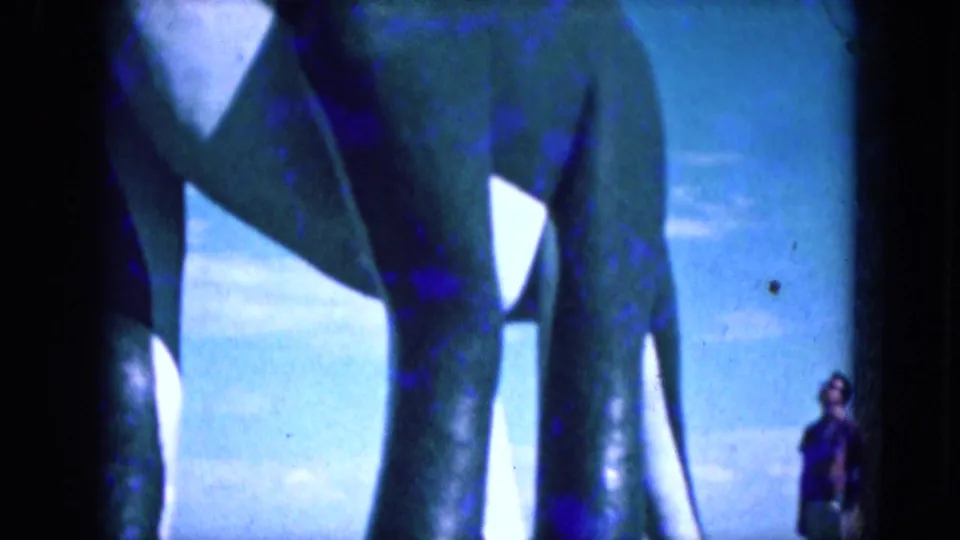 Shaky 1969 Lake Canyon, Arizona 8mm view of a large green dinosaur-like prop, showing legs, body sections, white accents, and blue sky.